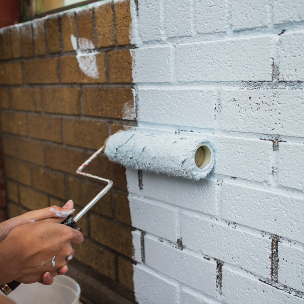 A person using a roller brush to apply fresh white paint to an exterior brick wall