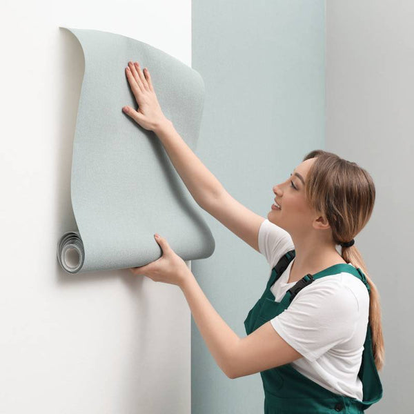 woman wearing overalls applying light green wallpaper to a white wall